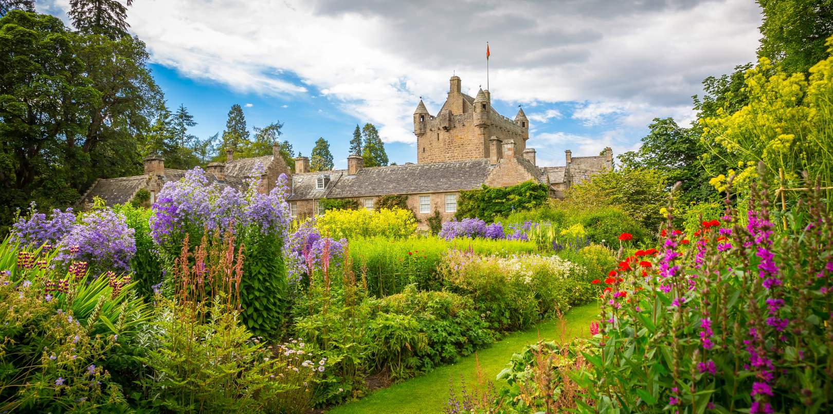Cawdor Castle's sandstone tower and slate roofs rising above colourful flower beds, lawn paths and mature trees under a blue, cloud-flecked sky